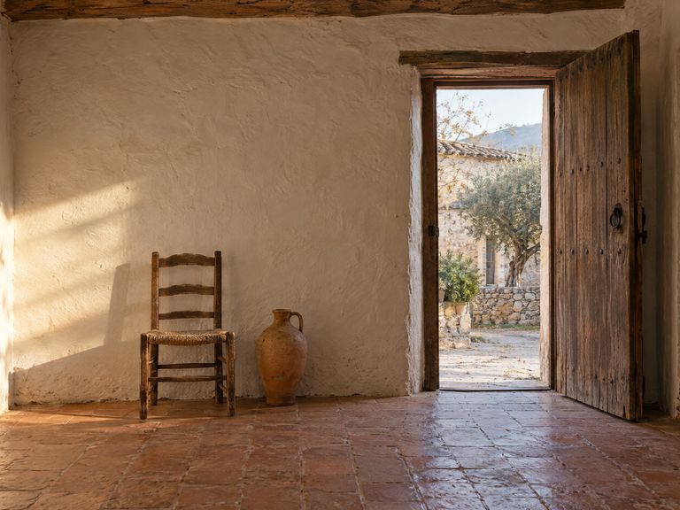 Entrada de una casa de pueblo con suelo de barro, una silla de anea y un cántaro junto a la pared; la puerta abierta deja ver un olivo.